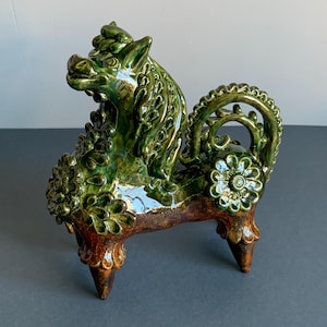May include: A ceramic sculpture of a mythical creature, featuring a green glaze with brown accents. The detailed design includes floral patterns and a decorative tail. The sculpture is mounted on four legs.