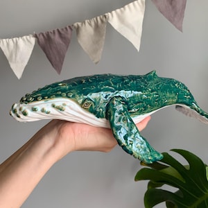 Puede incluir: Una figura de ballena jorobada de cerámica con un acabado verde oscuro brillante en el dorso y una parte inferior blanca lisa. La escultura muestra protuberancias texturizadas detalladas a lo largo de la cabeza y las aletas, capturando su forma.