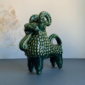 May include: A glossy, dark green ceramic figurine of a mythical creature with curled horns, a textured, scale-like body, and a long, protruding tongue. The four-legged animal stands upright, featuring a curved tail and a detailed, sculpted head.