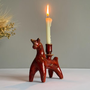 Puede incluir: Un portavelas rojo de cerámica con forma de caballo con una vela blanca encendida. El portavelas tiene un acabado brillante y un diseño rústico. La vela es alta y delgada, con una llama brillante.