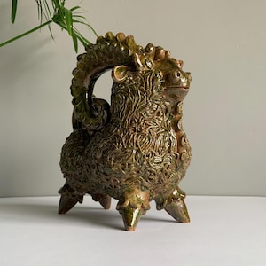 Op de afbeelding: Een keramische sculptuur van een mythisch wezen in groen- en bruintinten. Het gedetailleerde stuk heeft een textuur en wordt ondersteund door drie poten. De sculptuur heeft een decoratief, open cirkelontwerp op de rug.