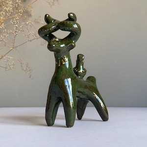 Puede incluir: Una escultura de cerámica de color verde oscuro brillante que representa una criatura de cuatro patas con grandes estructuras curvas en forma de cuernos en la cabeza. La figura tiene un esmalte liso y reflectante y una pequeña protuberancia.