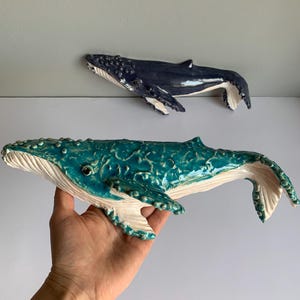 Puede incluir: Dos esculturas de ballenas de cerámica. Una es de color azul oscuro y la otra turquesa. La ballena turquesa se sostiene en una mano. Ambas ballenas tienen partes inferiores blancas y superficies texturizadas, con detalles como aletas y ojos.