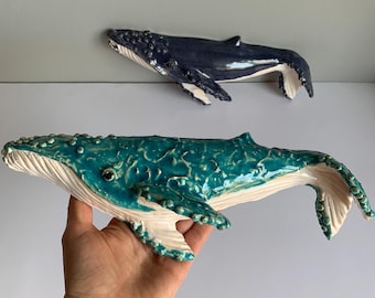 May include: Two ceramic whale sculptures. One is a dark blue color, and the other is a teal color. The teal whale is held in a hand. Both whales have white undersides and textured surfaces, with details like fins and eyes.
