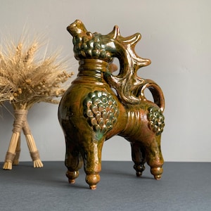 May include: A glazed ceramic figurine of a mythical creature with four legs, a long neck, and prominent antlers. The sculpture features a mottled brown and green finish with raised, scale-like textures on the body and neck, standing on four feet.