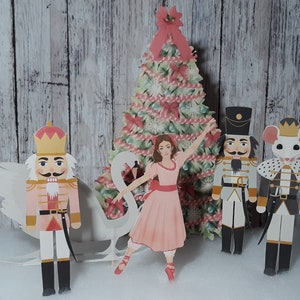 May include: A papercraft Christmas scene featuring a ballerina in a pink dress dancing with a white swan. The scene also includes a nutcracker soldier in a blue and gold uniform, a mouse king in a black and white uniform, and a Christmas tree decorated with red and white candy canes.