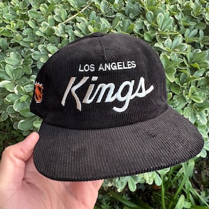 以下が含まれることがあります： 黒色のコーデュロイ野球帽。白の筆記体で「LOS ANGELES Kings」と書かれています。キャップの側面にはロサンゼルス・キングスのロゴが入っています。帽子にはカーブしたつばが付いています。