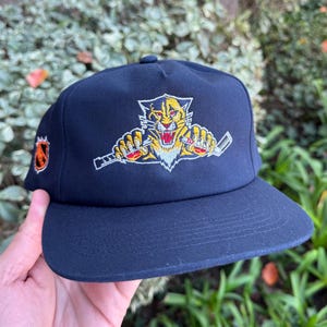 Peut inclure: Casquette de baseball bleu marine avec un logo brodé d'une panthère jaune et rouge rugissante tenant des crosses de hockey. La casquette a une visière plate et un petit logo d'équipe sur le côté.