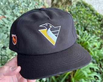 Può includere: Un cappellino da baseball nero con il logo ricamato dei Pittsburgh Penguins sul pannello frontale. Il logo raffigura un pinguino stilizzato in nero, bianco e giallo. Un piccolo logo arancione e bianco della NHL è ricamato sul lato destro.