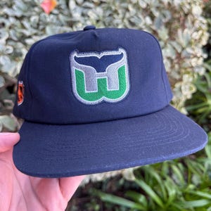 Könnte beinhalten: Marineblaue Baseballkappe mit flachem Schirm. Die Kappe zeigt ein Logo mit einer Walflosse und dem Buchstaben "W" in Grün und Silber. Ein orangefarbenes Logo befindet sich an der Seite. Ein Accessoire für Sportfans.