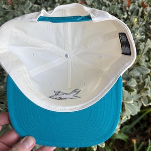 May include: A white baseball cap with a teal brim and adjustable strap. The underside of the cap is white with a shark logo. The cap is held by a hand.