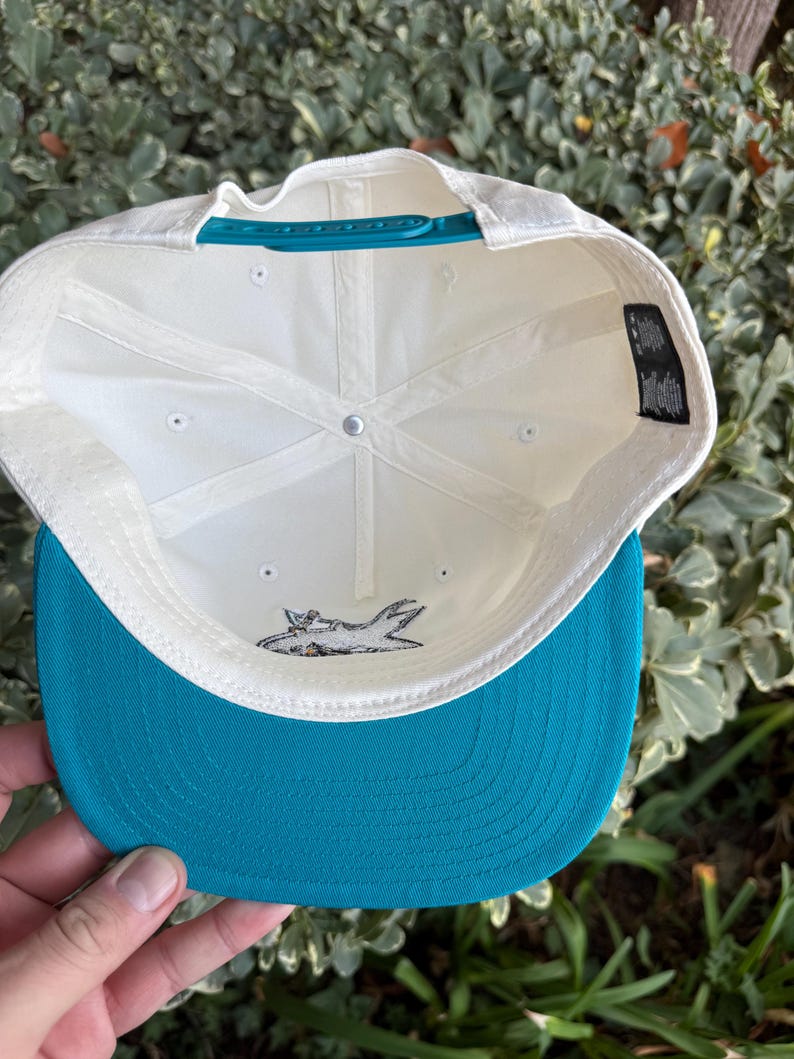 May include: A white baseball cap with a teal brim and adjustable strap. The underside of the cap is white with a shark logo. The cap is held by a hand.