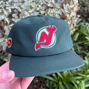 May include: A dark green baseball cap featuring the New Jersey Devils logo in red, white, and green. The logo is on the front of the cap, with a smaller logo on the side. The cap has a flat brim.