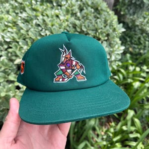 May include: A green baseball cap with a team logo on the front. The logo is a stylized coyote in shades of purple, brown, and green. The cap has a curved peak and a structured crown. The cap is a solid green colour.