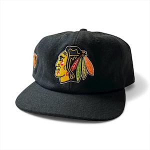 May include: Black baseball cap with a flat peak. The cap features an embroidered logo of a Native American chief in yellow, red, and green. The cap is made of a textured fabric and has a curved peak.