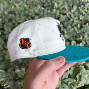 May include: A white baseball cap with a teal brim. The cap features a team logo on the front and a team logo patch on the side. The cap is held by a hand.