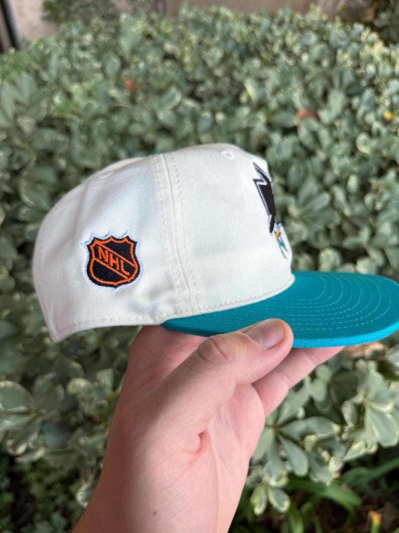 May include: A white baseball cap with a teal brim. The cap features a team logo on the front and a team logo patch on the side. The cap is held by a hand.