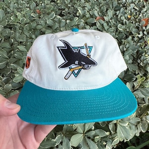 Puede incluir: Una gorra de béisbol blanca y verde azulado con el logotipo de los San Jose Sharks. La gorra tiene una visera verde azulado y un botón verde azulado en la parte superior. El logotipo es un tiburón negro con palos de hockey y un triángulo.