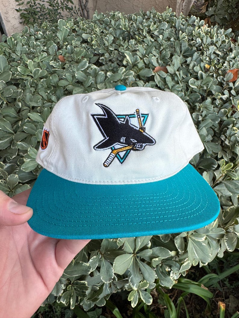 May include: A white and teal baseball cap featuring the San Jose Sharks logo. The cap has a teal brim and a teal button on top. The logo is a black shark with hockey sticks and a triangle.