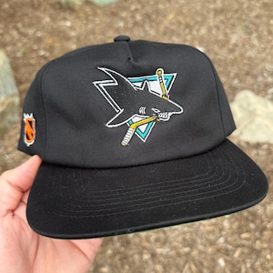May include: Black baseball cap featuring the San Jose Sharks logo. The logo includes a shark, hockey stick, and a triangle. The cap has a flat brim and a green underside. An orange and black logo is on the side.