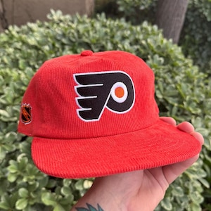 Könnte beinhalten: Rote Cord-Baseballkappe mit dem Logo der Philadelphia Flyers in Schwarz, Weiß und Orange. Die Kappe hat einen gebogenen Schirm und ein kleines Emblem an der Seite. Die Kappe wird in einer Hand gehalten.