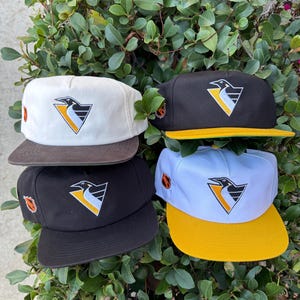 Könnte beinhalten: Vier Baseballkappen mit flachem Schirm und Snapback-Verschluss. Die Kappen sind weiß, schwarz und gelb und tragen ein Pinguin-Logo in einem Dreieck. Die Kappen haben braune, gelbe und grüne Schirme.