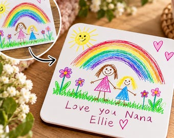 May include: A square coaster featuring a colorful child's drawing of a yellow sun, a rainbow, two figures, flowers, and hearts. The text reads Love you Nana Ellie with a heart symbol. The design is printed on a white surface with rounded corners.