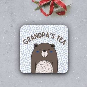 May include: A white coaster with a blue and white polka dot pattern. The text "Grandpa's Tea" is written in black letters. A brown bear with a white belly and blue eyes is in the center of the coaster.