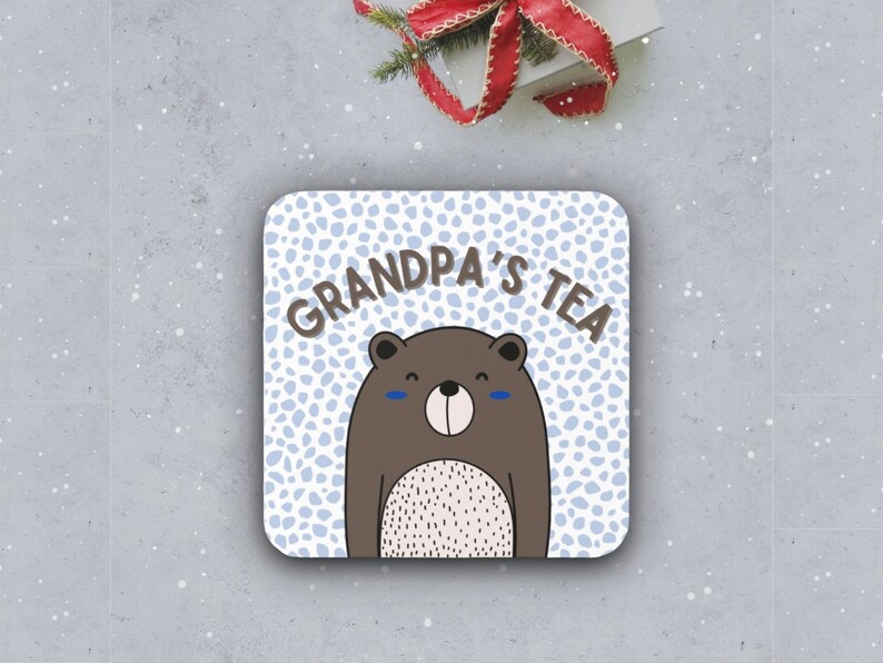 May include: A white coaster with a blue and white polka dot pattern. The text "Grandpa's Tea" is written in black letters. A brown bear with a white belly and blue eyes is in the center of the coaster.