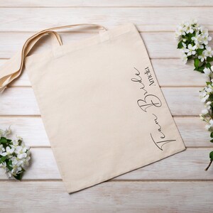 May include: A beige canvas tote bag with long handles, featuring the text "Team Bride" in black script. The bag is lying on a light wood surface, decorated with sprigs of white flowers.