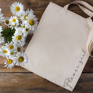 May include: A cream-colored tote bag with the word "Flower Girl" in a cursive font. The bag is next to a bouquet of daisies with white petals and yellow centers. The background is a rustic wooden surface.