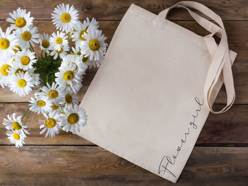 May include: A cream-colored tote bag with the word "Flower Girl" in a cursive font. The bag is next to a bouquet of daisies with white petals and yellow centers. The background is a rustic wooden surface.