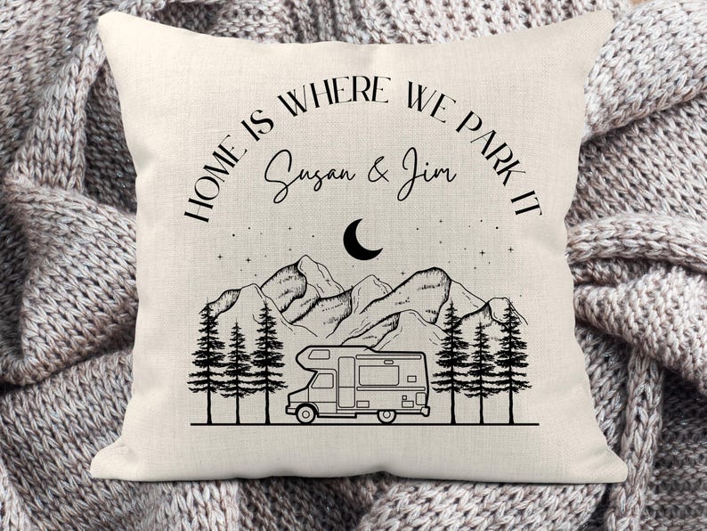 May include: A square off-white throw pillow featuring a black line art graphic of a camper van, pine trees, mountains, a crescent moon, and stars. The text reads "HOME IS WHERE WE PARK IT" above the names "Susan & Jim" in black ink.