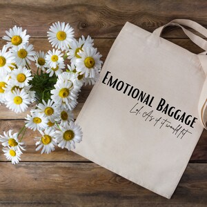 May include: A white canvas tote bag with black text that reads "Emotional Baggage Lot: As if it would fit". The bag is lying on a wooden surface with a bouquet of white daisies in the upper left corner.