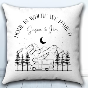 May include: A white square throw pillow featuring a black line art illustration of a camper van, pine trees, mountains, and a crescent moon. The text reads "HOME IS WHERE WE PARK IT" and the names "Susan & Jim" in black lettering.