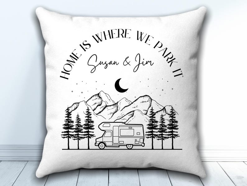 May include: A white square throw pillow featuring a black line art illustration of a camper van, pine trees, mountains, and a crescent moon. The text reads "HOME IS WHERE WE PARK IT" and the names "Susan & Jim" in black lettering.