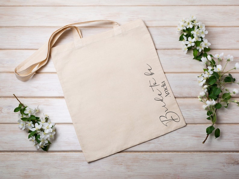 May include: A beige canvas tote bag with the words "Bride to be" and a name in black script. The bag has long handles and is lying on a light wood surface, with white flowers and green leaves.