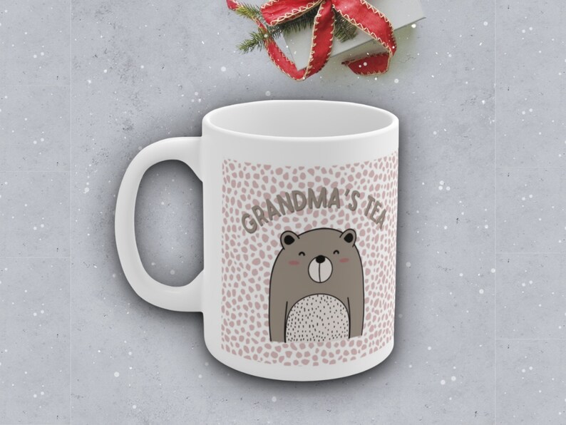 May include: White ceramic mug with a pink and white polka dot pattern. The mug has a brown bear illustration and the text "Grandma's Tea" in black.