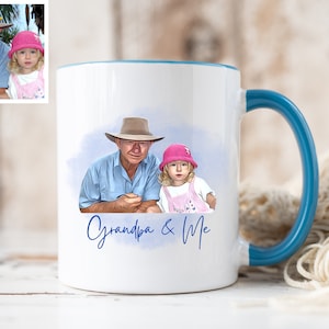 May include: A white ceramic mug with a blue handle and rim, featuring a digital illustration of an older person in a tan hat and a child in a pink hat. Below the illustration, the text Grandpa & Me is written in blue script.