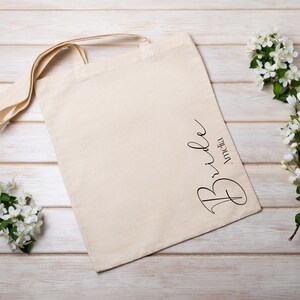 May include: A cream-colored tote bag with the word "Bride" and the name "Amelia" printed in black script. The bag has long handles and is placed on a light wood surface, with white flowers and green leaves.
