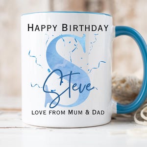 May include: White ceramic mug with a blue handle and rim. The mug features the text "HAPPY BIRTHDAY" above a large blue letter "S" with the name "Steve" below it. The text "LOVE FROM MUM & DAD" is at the bottom.