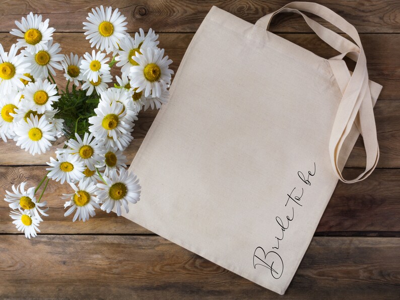 May include: A cream-colored tote bag with the words "Bride to be" in elegant script. The bag has long straps and is next to a bouquet of white daisies with yellow centers. The background is a rustic wooden surface.