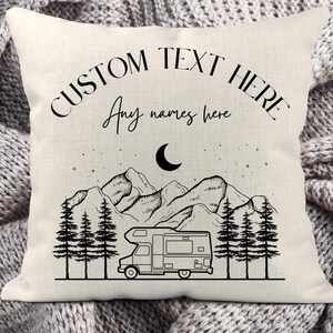 May include: A square off-white throw pillow featuring a black line art illustration of a camper van, pine trees, mountains, and a crescent moon. The text CUSTOM TEXT HERE and Any names here is printed in black ink on the upper portion.