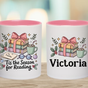 May include: White ceramic mug with a pink interior and handle, featuring a festive design. The design includes a stack of books tied with a pink bow, a mug of cocoa, and the text "'Tis the Season for Reading".