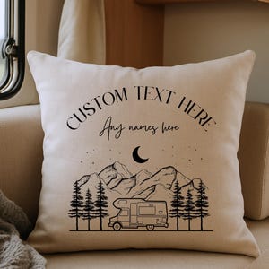 May include: A square off-white throw pillow featuring a black line art illustration of a camper van, pine trees, mountains, and a crescent moon. The text reads CUSTOM TEXT HERE and Any names here in a mix of serif and script fonts.