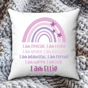 May include: A white cushion with a rainbow design in shades of pink, purple, and white. The rainbow has stars on top. The cushion has the text "I am special. I am Loved I am brave. I am enoug I am beautiful. I am Strong I am happy. I belong I am Ellie" printed on it.