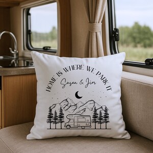 May include: A white square throw pillow featuring a black line art graphic of a camper van, pine trees, mountains, stars, and a crescent moon. The text reads "HOME IS WHERE WE PARK IT" above the names "Susan & Jim" in a script font.