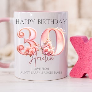 May include: White ceramic mug with a pink handle and interior, featuring the text "HAPPY BIRTHDAY 30 Amelia" with floral accents. The mug is personalized with a name and a message from family. A pink "X" shape is next to the mug.