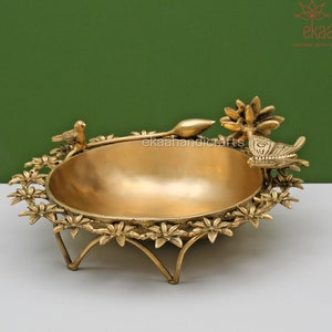 Könnte beinhalten: Eine goldene Metallschale mit einem dekorativen Blumendesign am Rand. Die Schale steht auf einem Ständer mit drei Beinen und zeigt ein Vogel- und Schmetterlingsdesign.