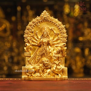 May include: A gold-coloured metal statue of a Hindu goddess with multiple arms, sitting on a throne with two lions at her feet. The statue is intricately detailed and has a traditional design.